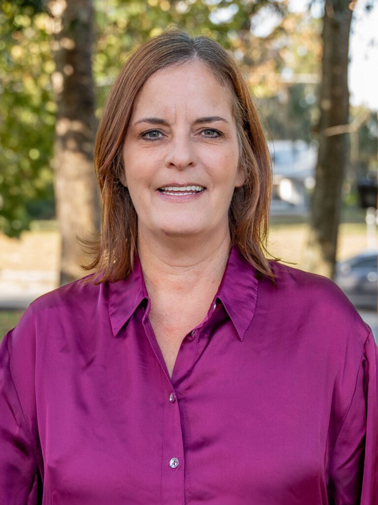 Smiling woman in a purple blouse outdoors with trees in the background.